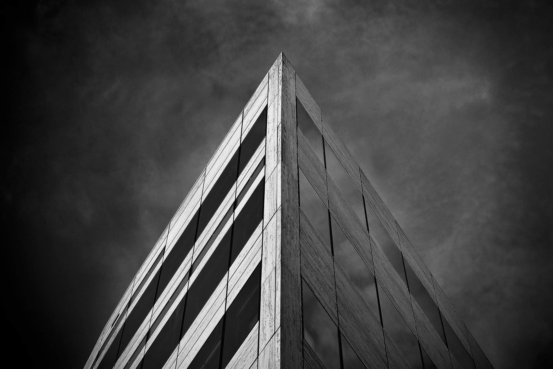 Black and white low-angle photo of the corner of a multi-storey glass and concrete building against a moody sky
